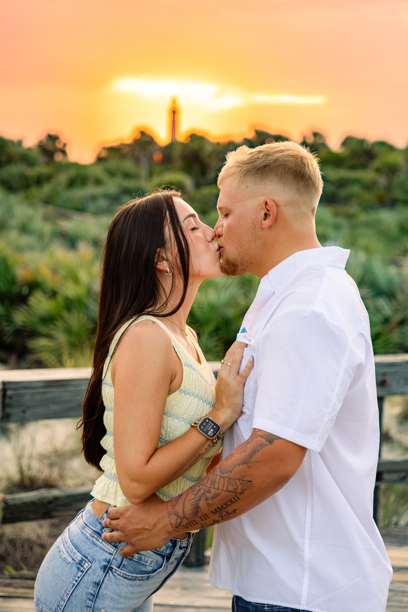Couple sharing a romantic kiss at sunset with a lighthouse silhouette and orange sky backdrop.
