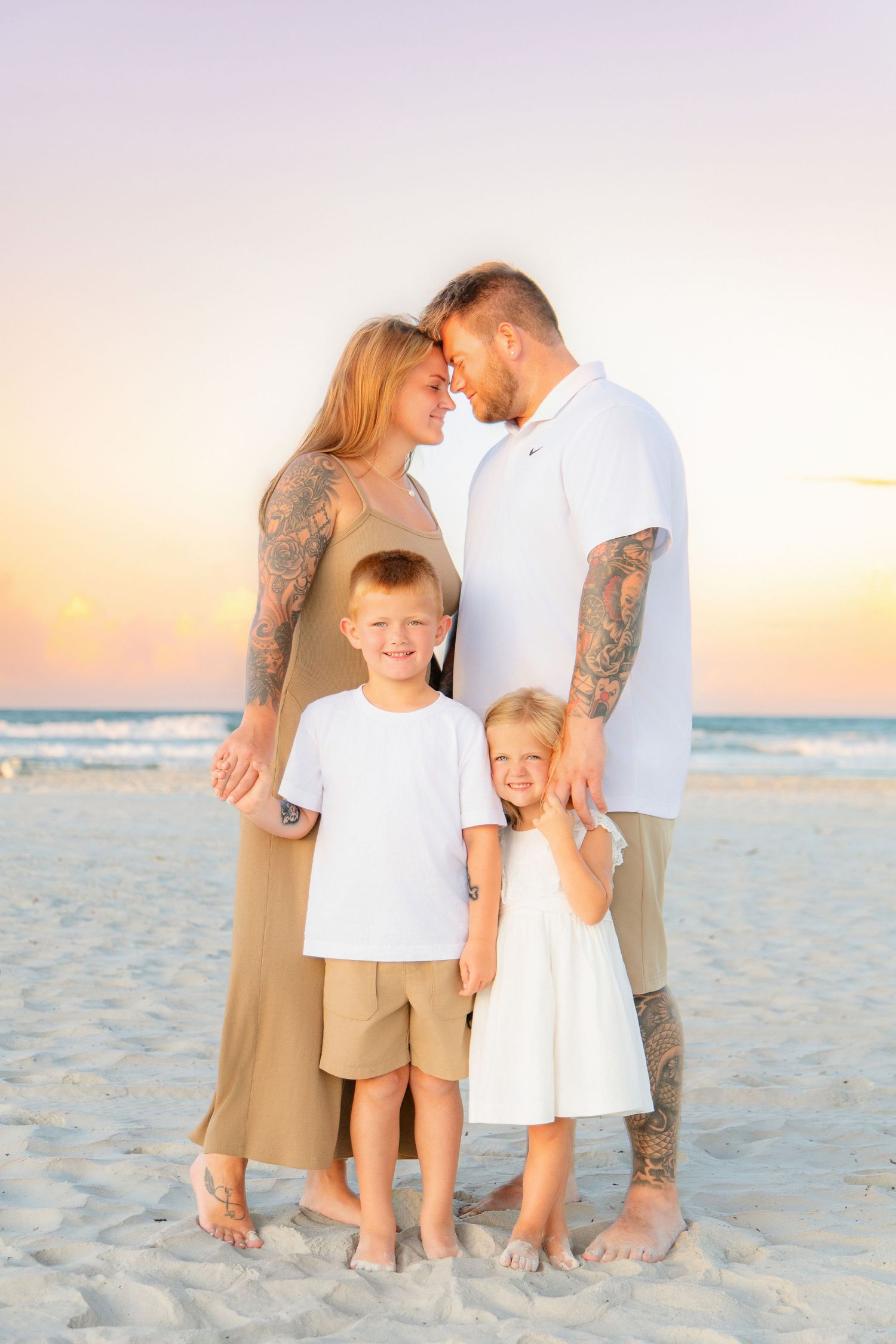 Family of four poses on sandy beach at sunset, parents touching foreheads while children smile in matching outfits.