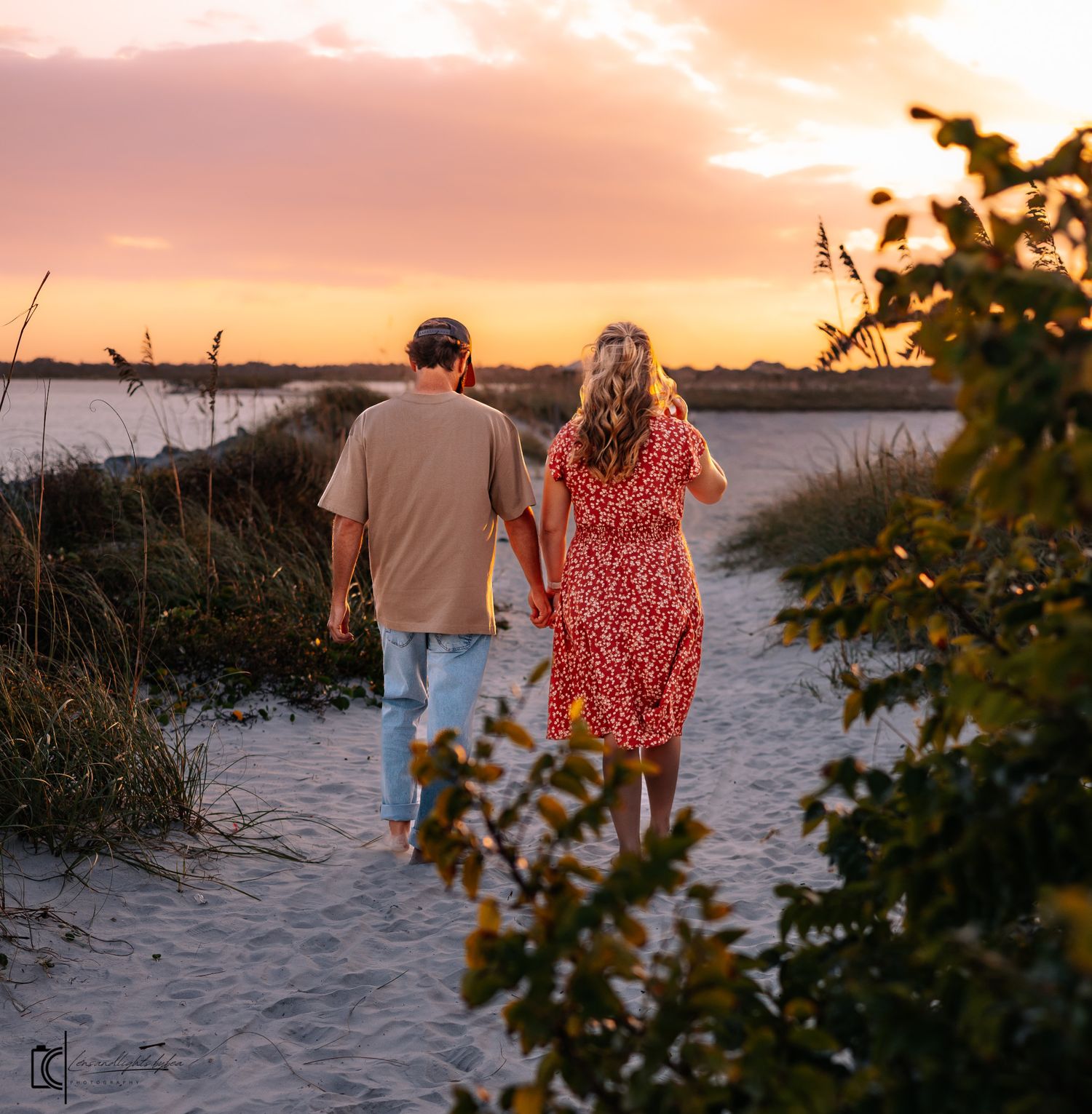 A couple walks along a sandy beach path at sunset with golden light filtering through coastal vegetation.