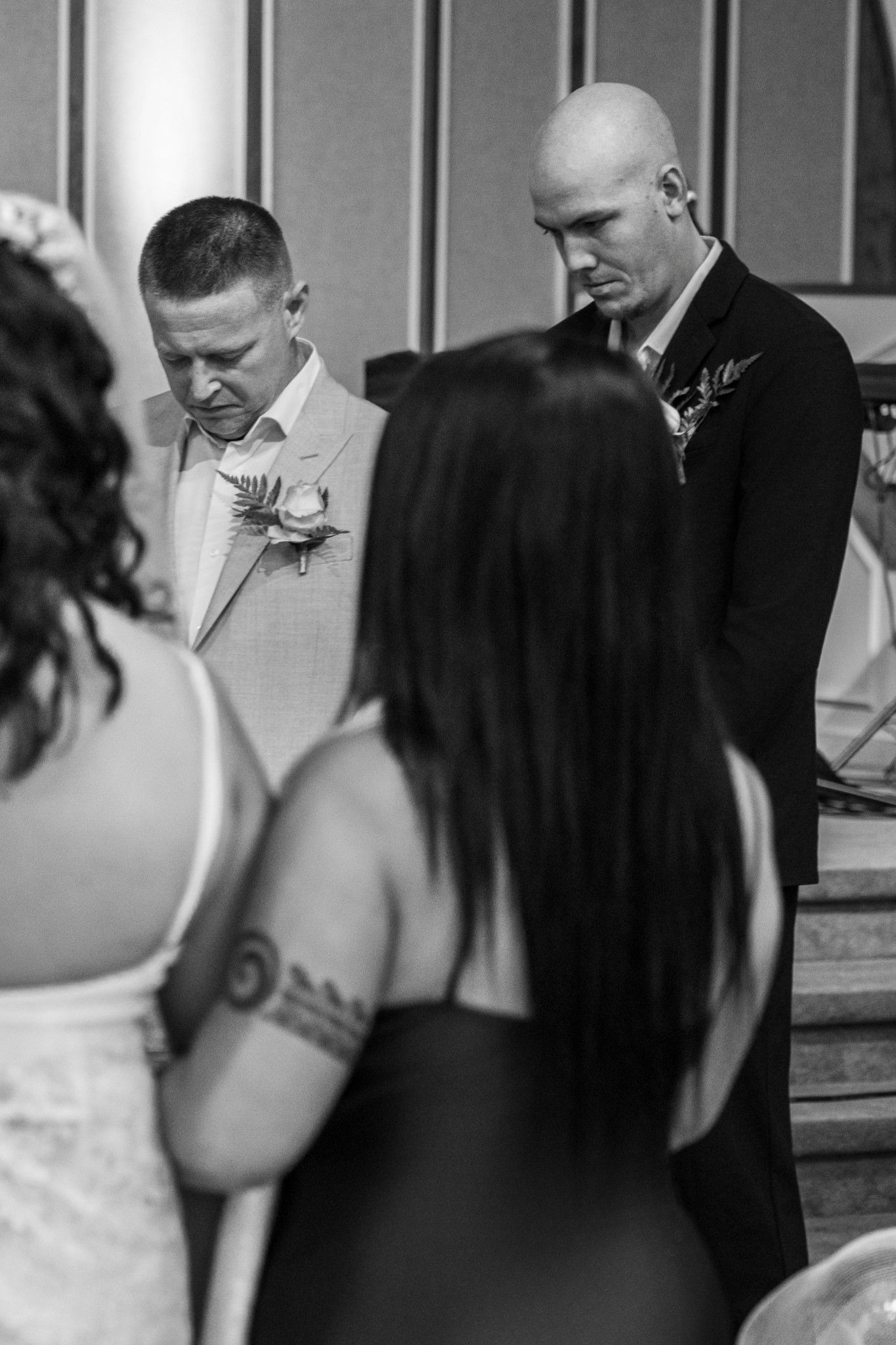 Black and white wedding ceremony photo of groom in light suit with boutonniere bowing head during prayer.