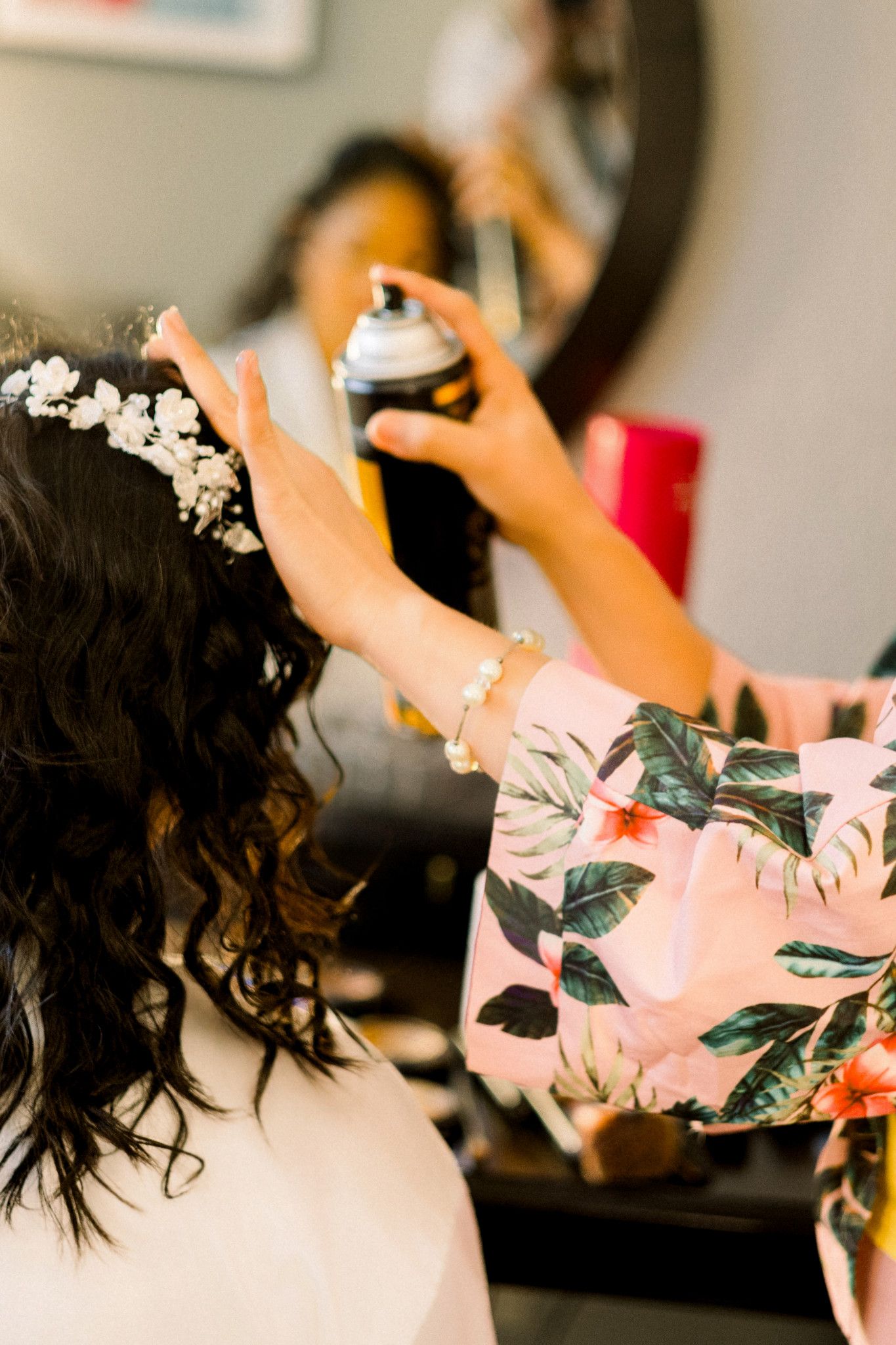 Hairstylist applying hairspray to bride's dark curly hair adorned with floral crown during wedding preparation.