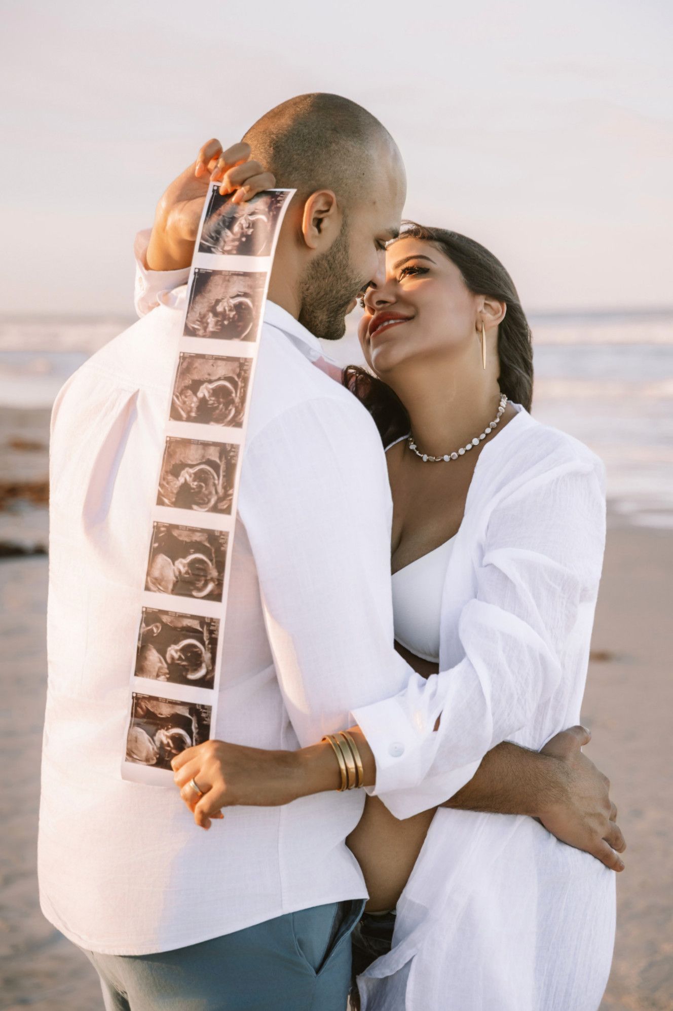 Couple embracing on beach at sunset, holding ultrasound photos, announcing pregnancy in matching white outfits.