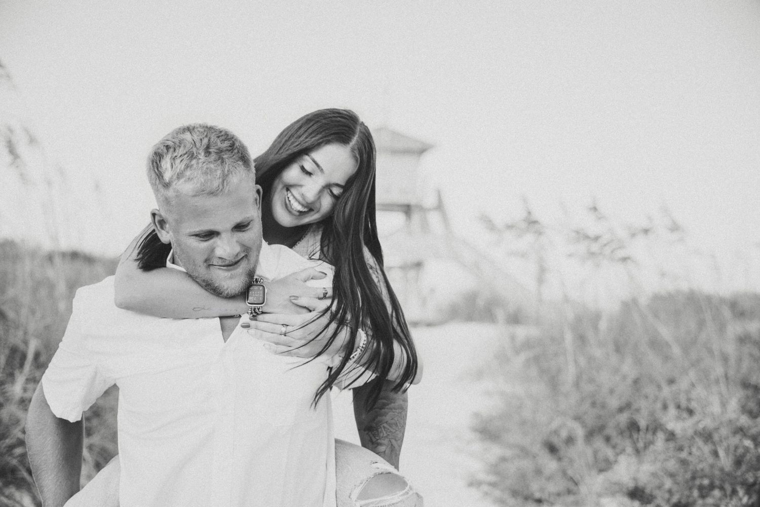 Black and white photo of a man giving a laughing woman a piggyback ride on a sandy beach path.
