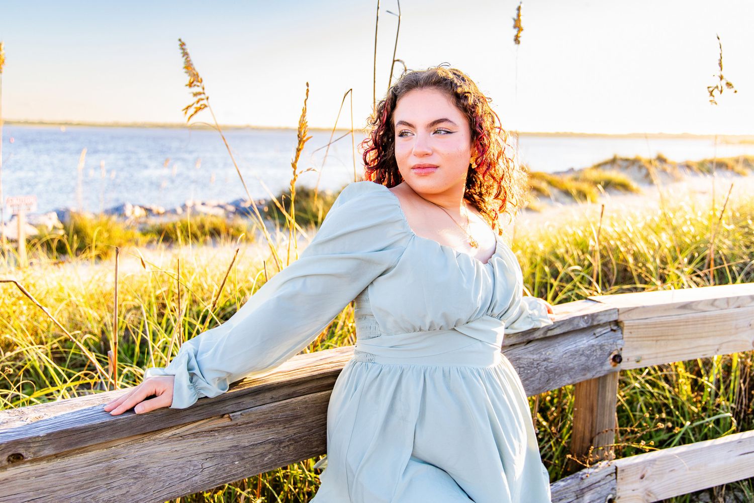 Young woman with curly hair in a light blue dress leaning on a wooden railing among beach grasses with the ocean behind her.
