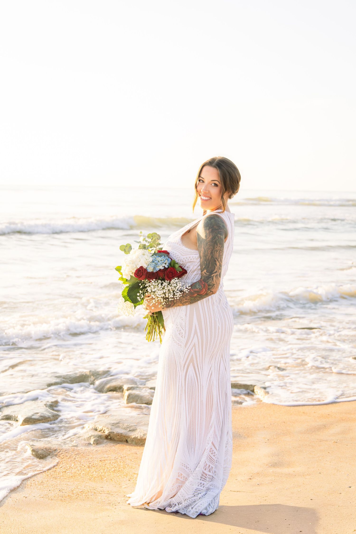 Tattooed bride in white lace gown holds red and white bouquet on a sunlit beach.