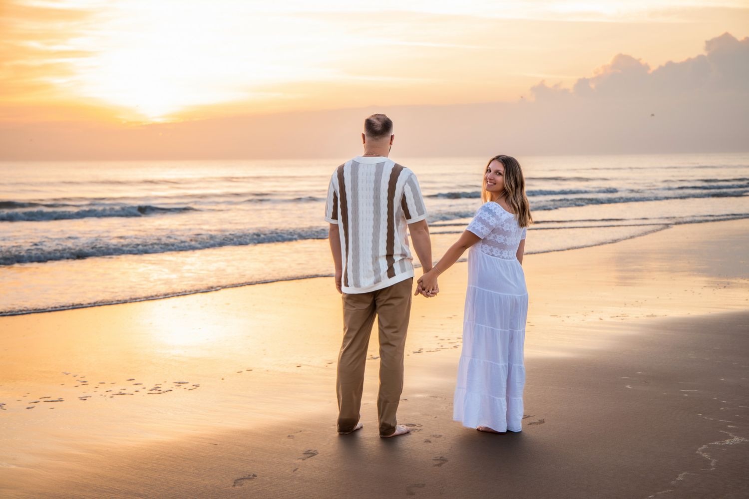 A couple holds hands on a golden beach at sunset as waves roll in along the shoreline.
