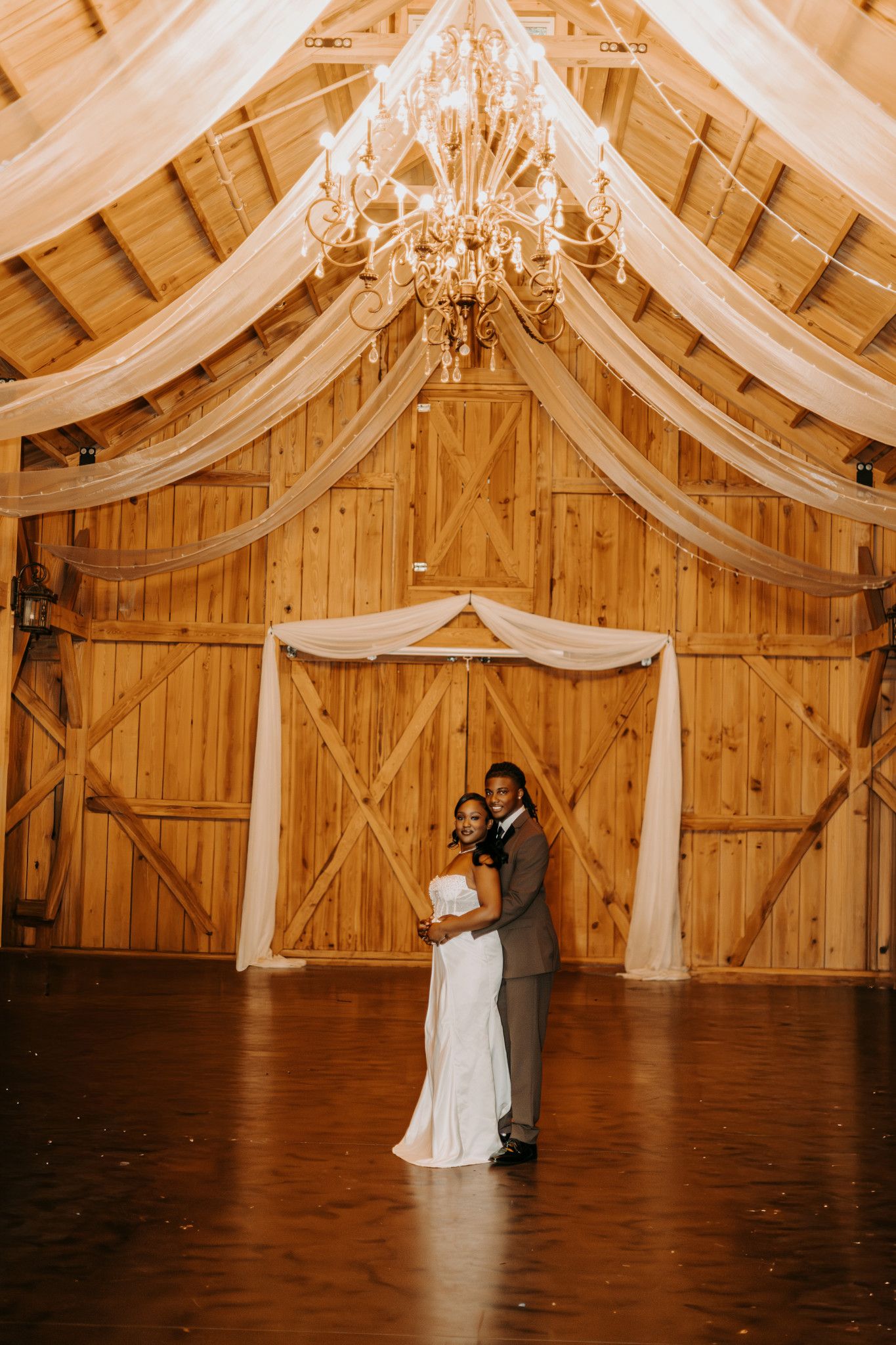 A romantic first dance in a rustic wooden barn venue decorated with elegant white draping and chandeliers.