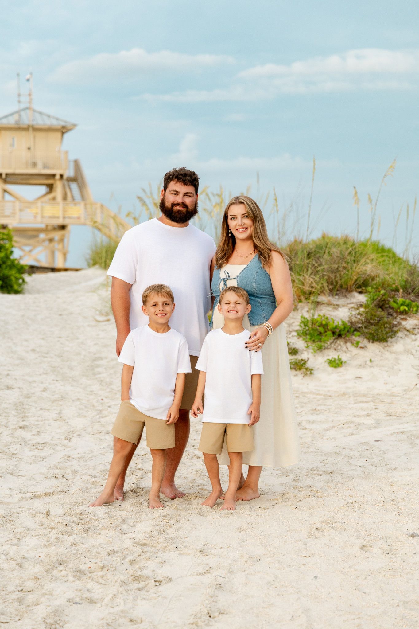Family of four posing on sandy beach path with lifeguard tower, wearing white shirts and khaki shorts on a sunny day.