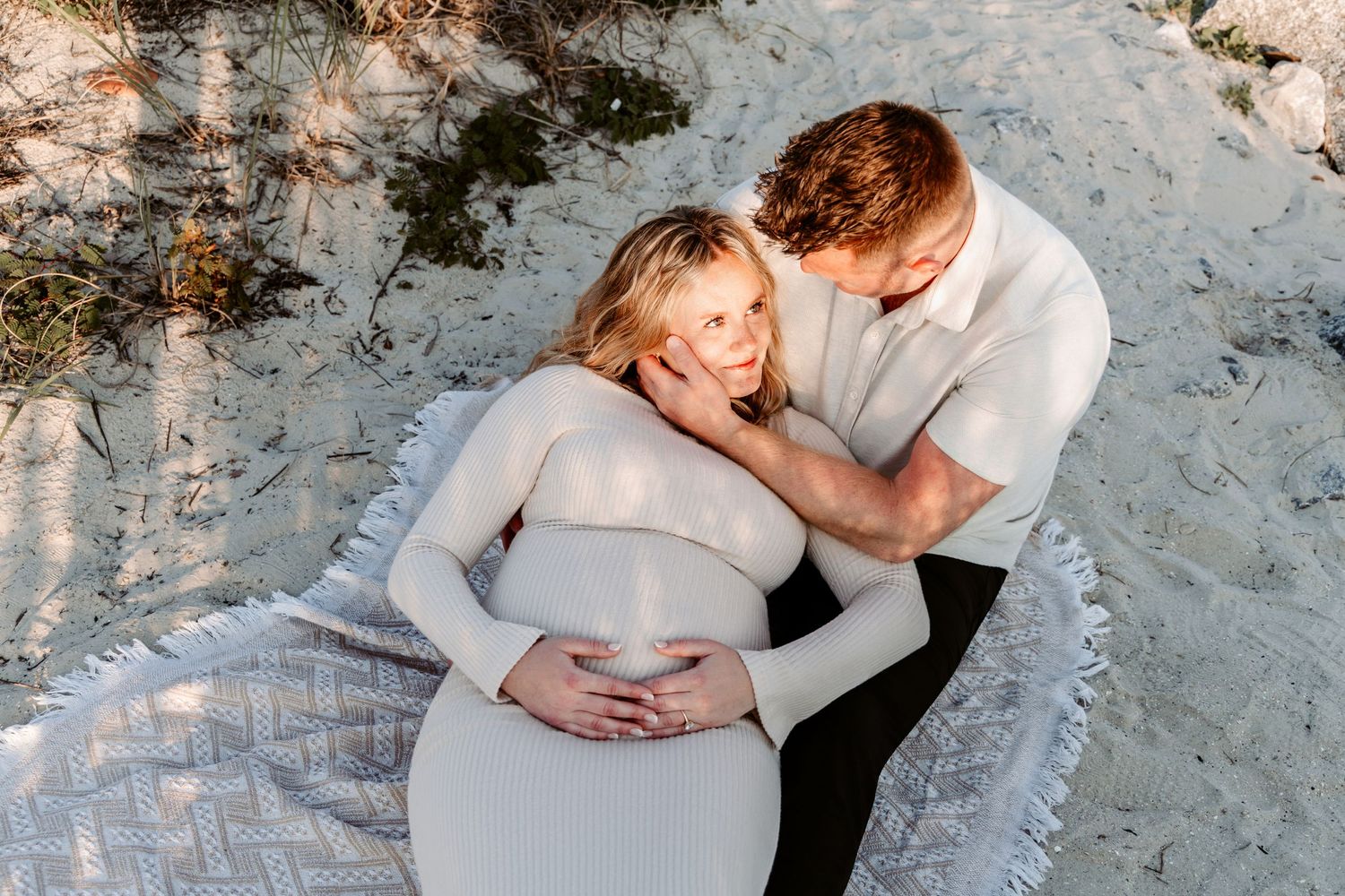 Couple sitting on beach blanket during maternity photoshoot, man tenderly holding pregnant womans face while she cradles her bump.