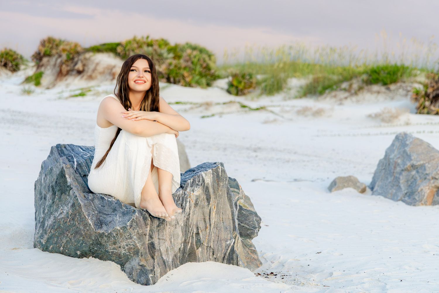 A young woman in a white dress sits on rocks at a sandy beach with dunes and green shrubs behind her.