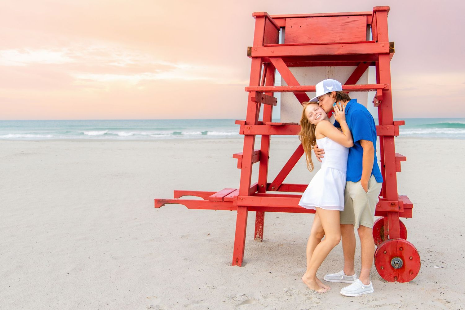 Couple embracing near a red lifeguard tower on a sandy beach at sunset.