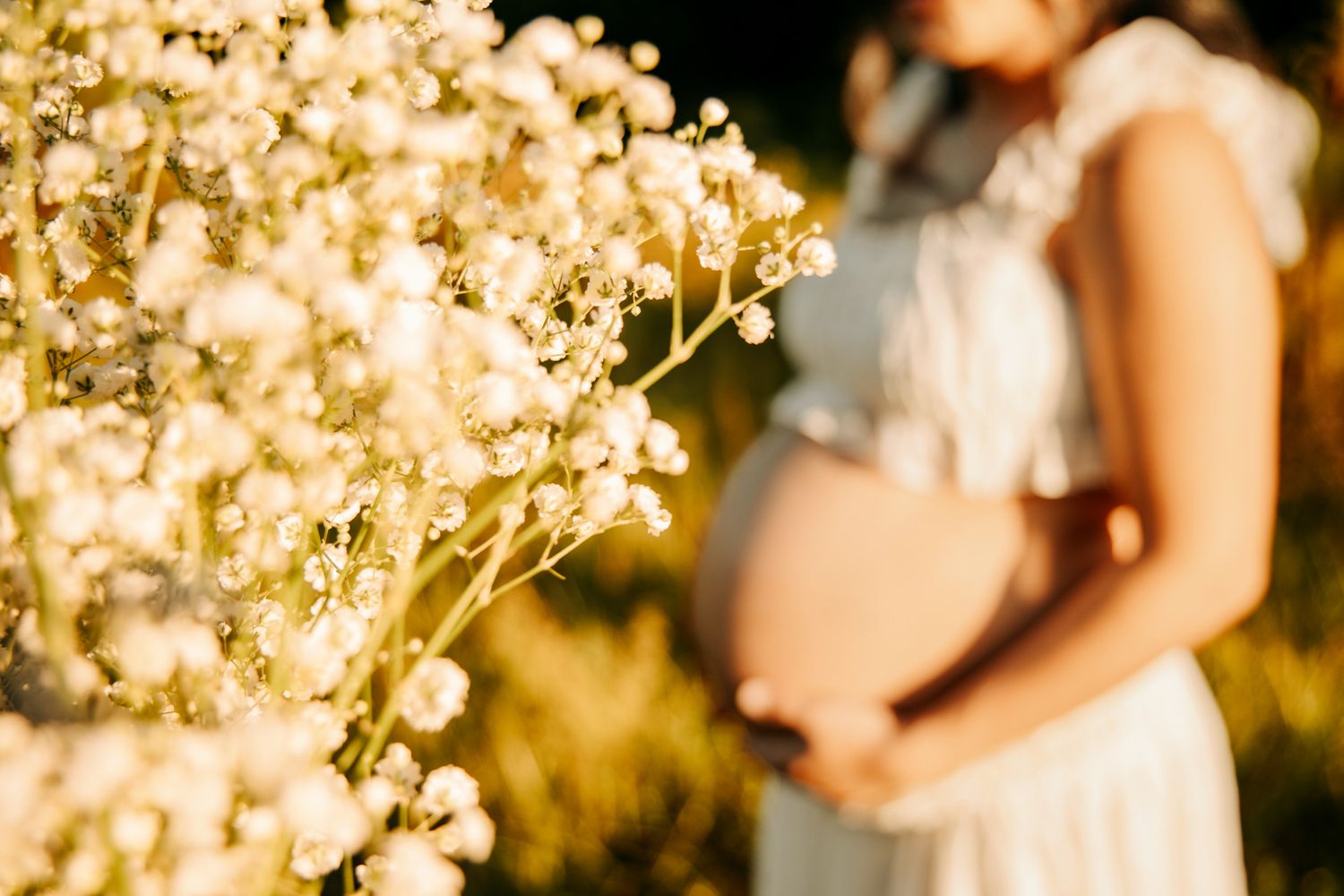 Pregnant woman in white dress cradling her baby bump outdoors near white wildflowers in warm golden sunlight.