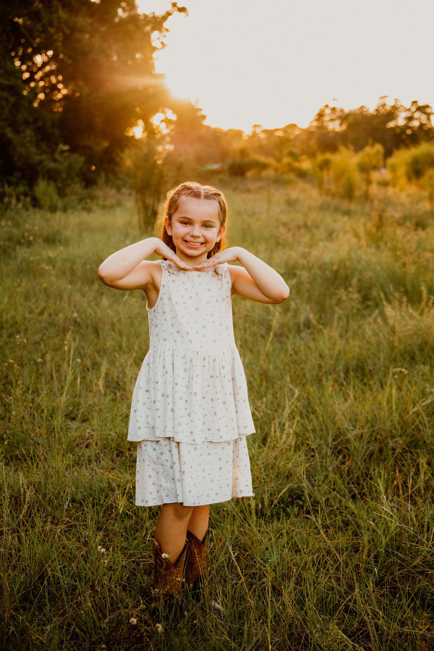 Young girl in white polka dot dress and cowboy boots smiling in a sunlit green field at golden hour.