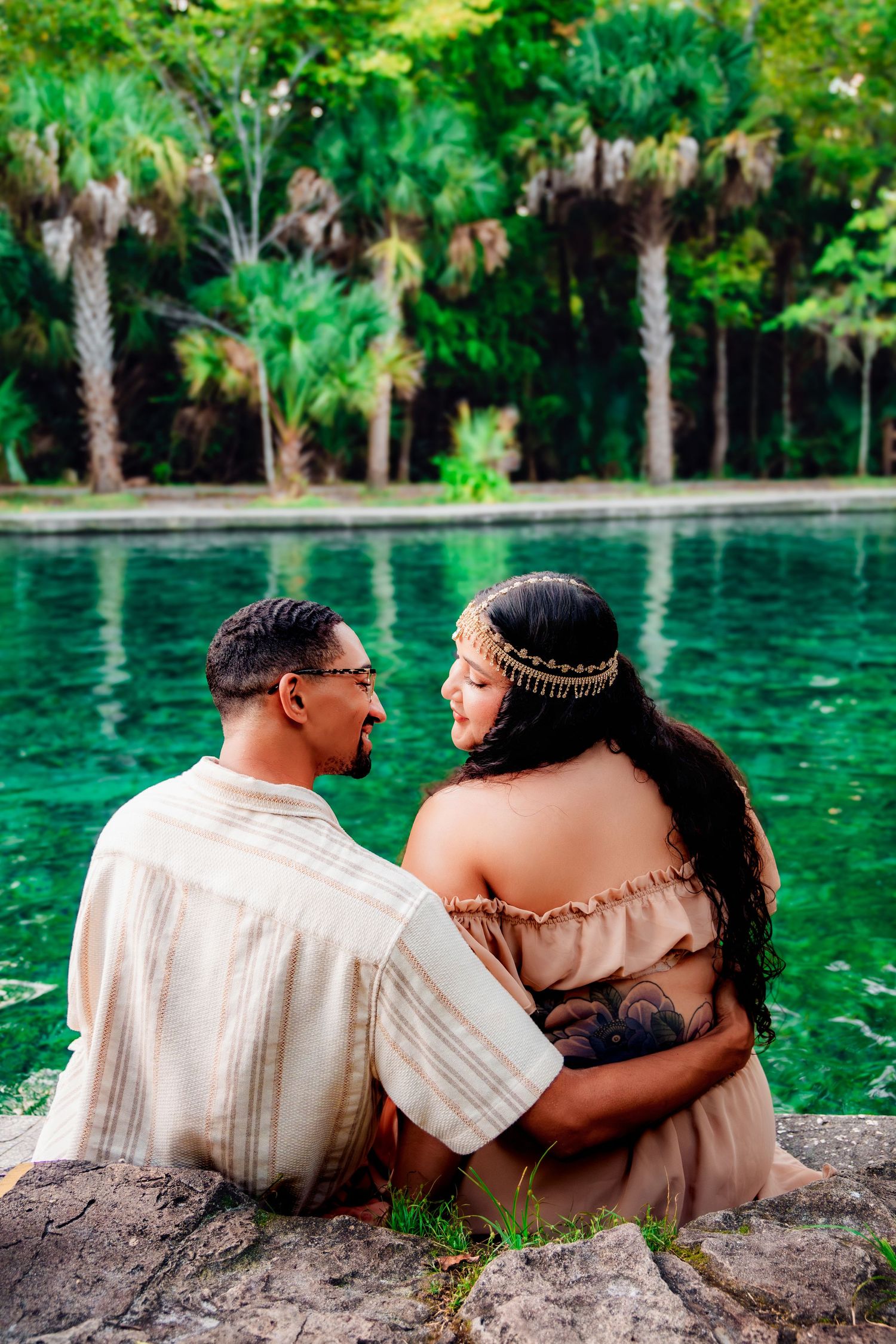 Couple sitting by a turquoise cenote surrounded by lush tropical palms, sharing a tender embrace in bohemian attire.