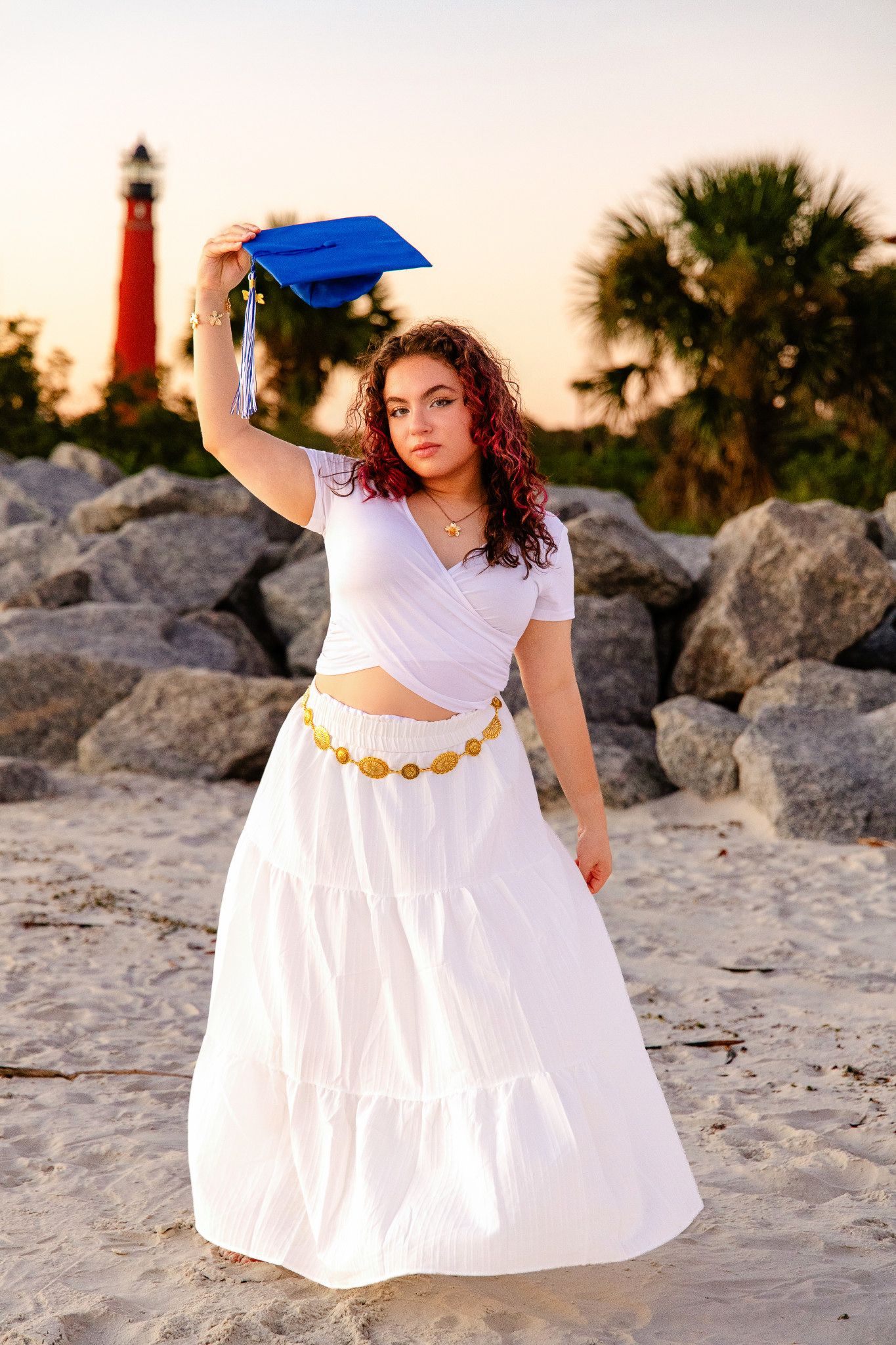 Graduating senior in a white outfit and gold belt holds a blue cap overhead on a sandy beach with a red lighthouse in the background.