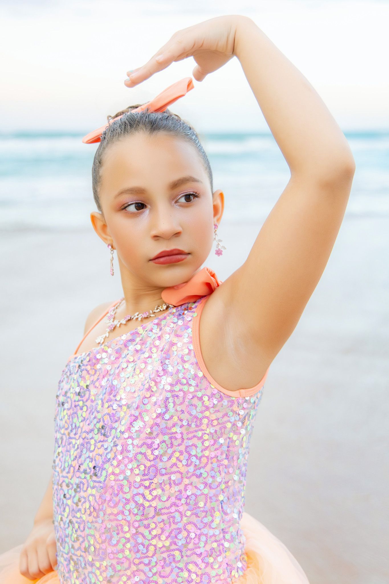 Young dancer in sequined pink dress poses gracefully on beach, arm raised overhead, wearing orange bow and jewelry.