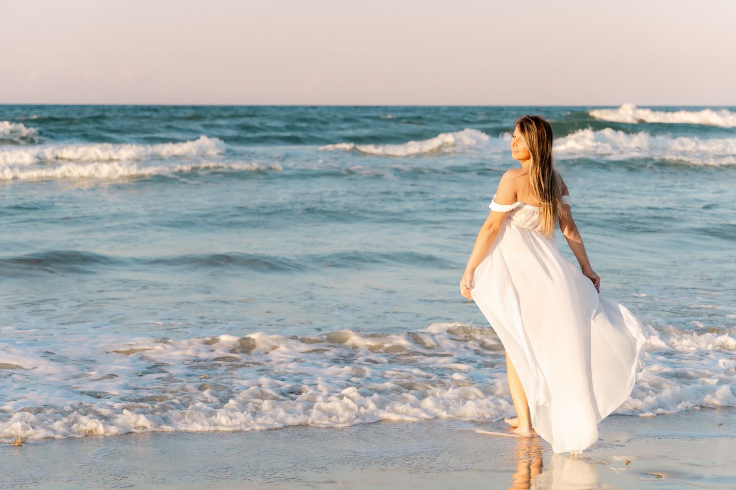 Woman in flowing white dress standing at ocean's edge on a sandy beach at sunset.