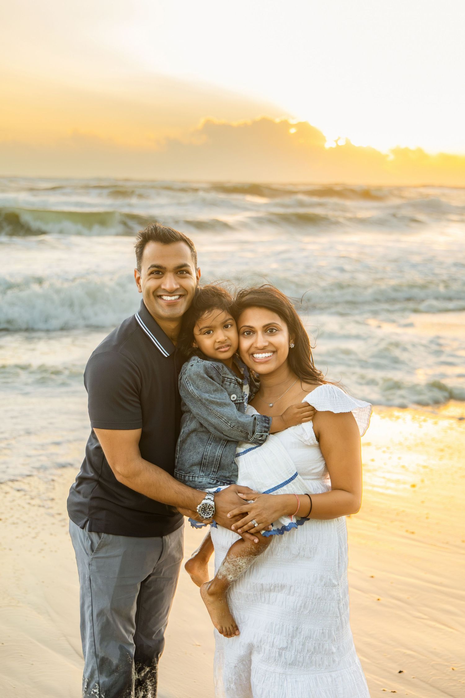 Smiling couple embracing their young child on a sandy beach at sunset with ocean waves and a golden sky behind them.