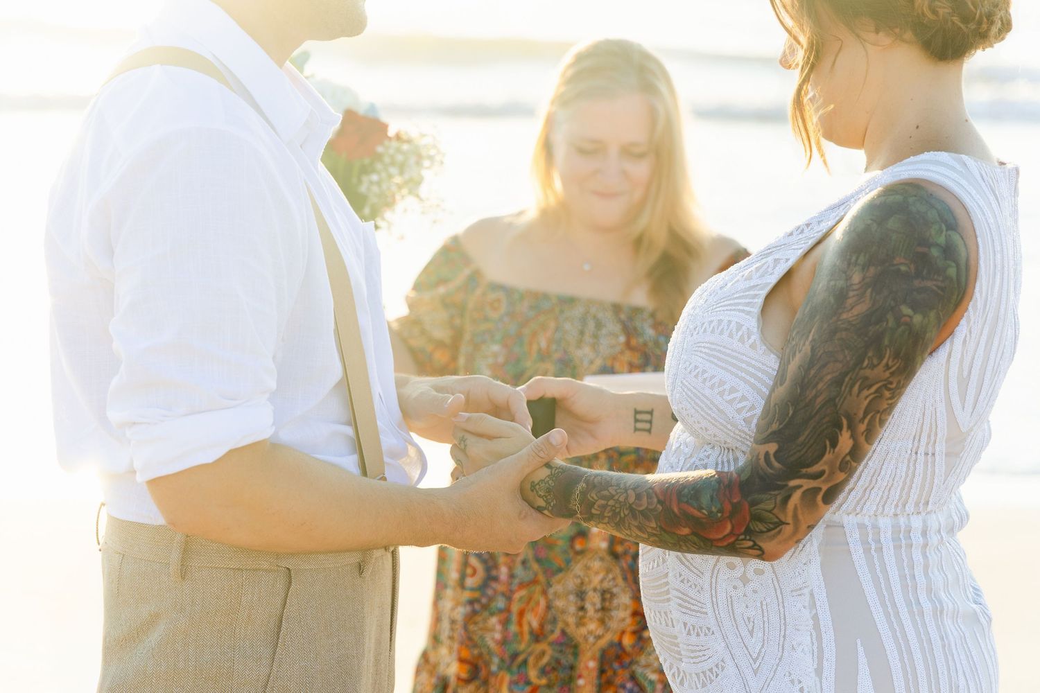 Couple exchanges rings at beach wedding ceremony with officiant in golden light.