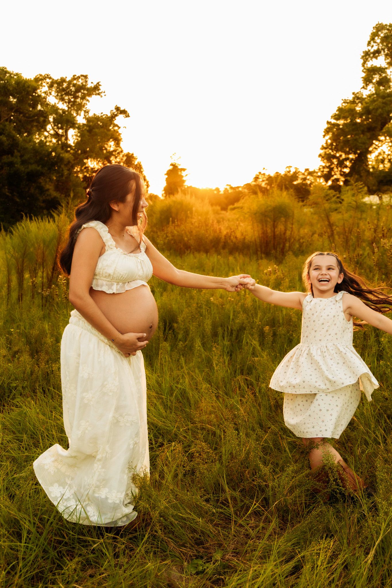 Laughing girl in white dress holds hands with pregnant mother in sunny green meadow.