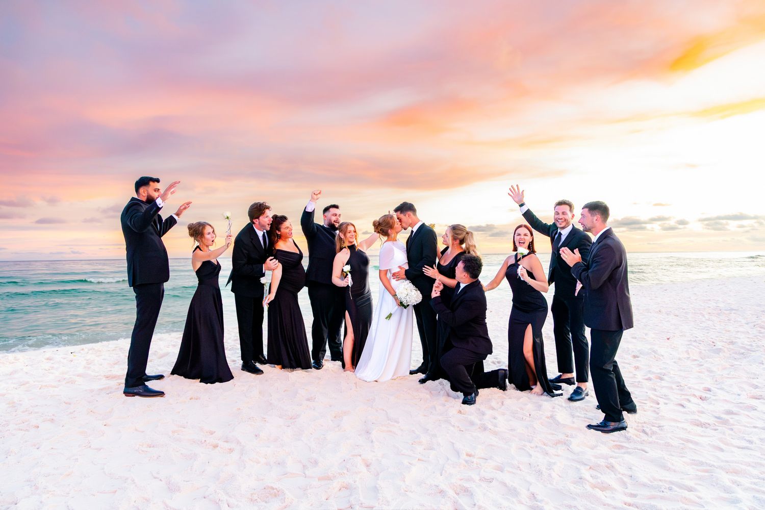 Wedding party celebrating on a beach at sunset, bride and groom kissing surrounded by guests in black formal attire.