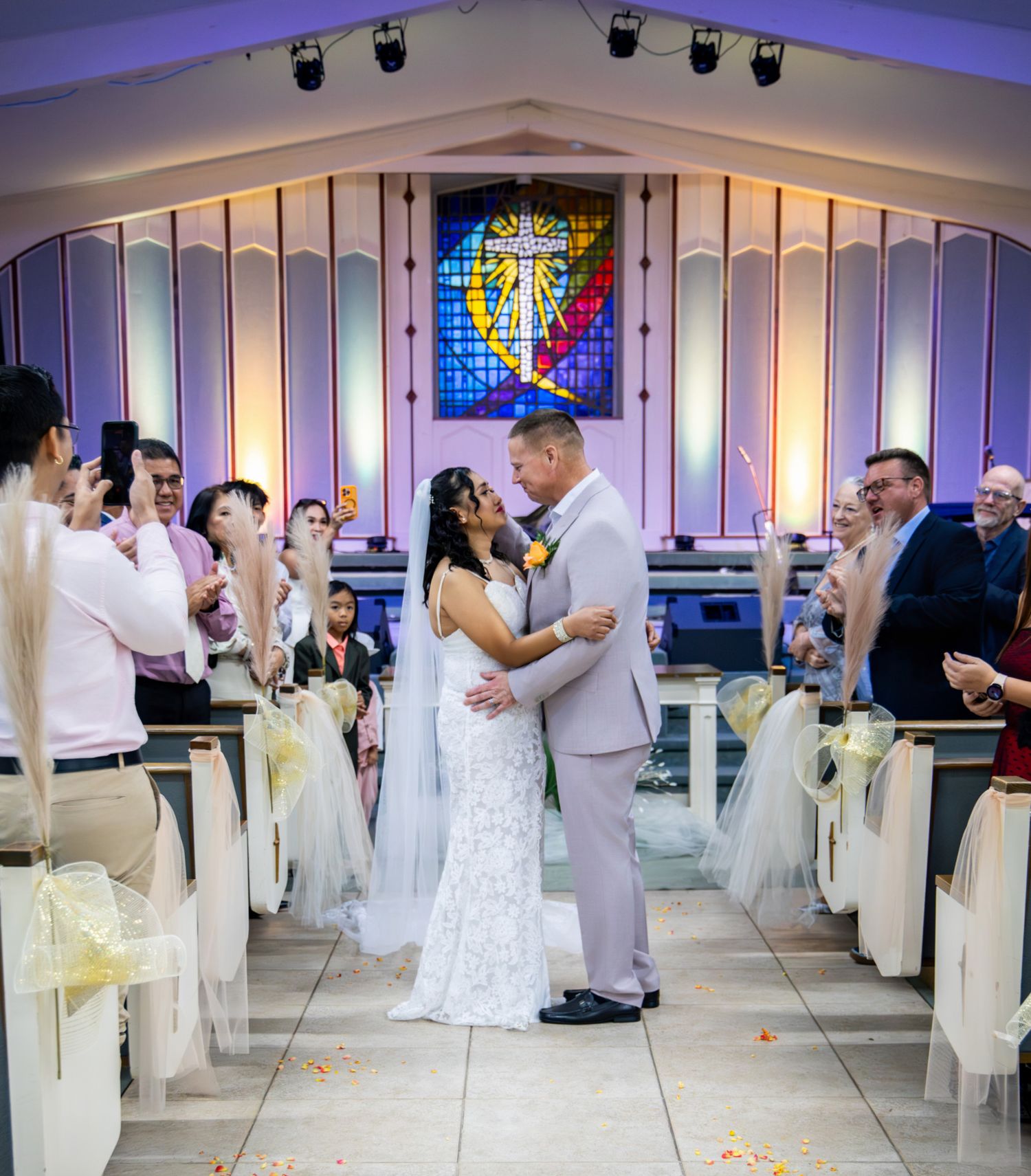Bride and groom share first kiss at chapel altar, surrounded by cheering guests and colorful stained glass window.