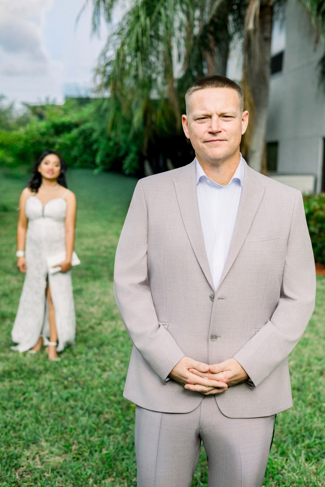Groom in light gray suit stands outdoors with bride in white dress visible in background at wedding.