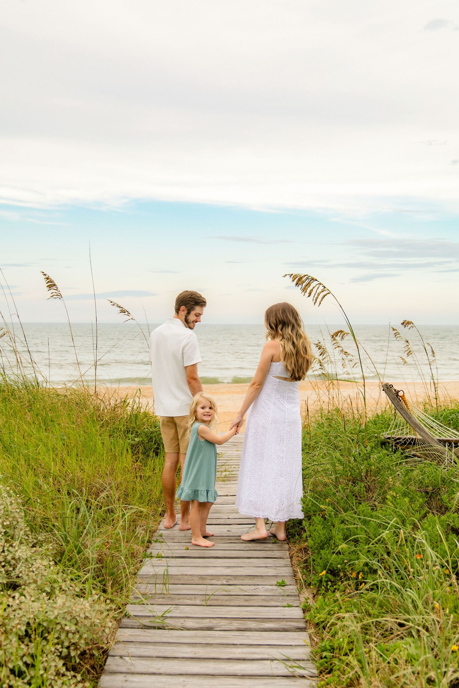 Family of three holding hands on a wooden beach boardwalk surrounded by sea oats, facing the ocean on a cloudy day.