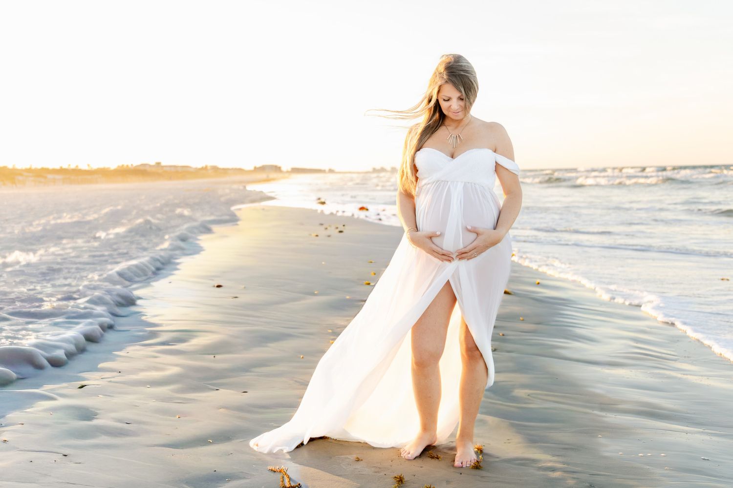 Pregnant woman in white off-shoulder gown standing on sandy beach at golden hour, cradling baby bump.