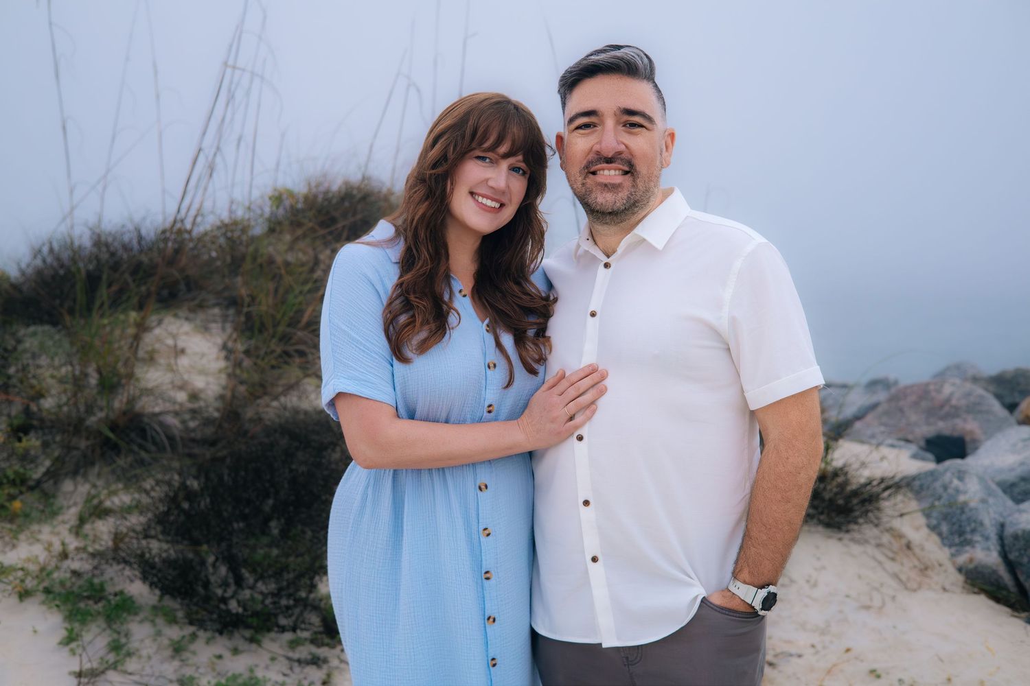 A couple in light blue and white outfits poses together for romantic engagement photos on a coastal beach.