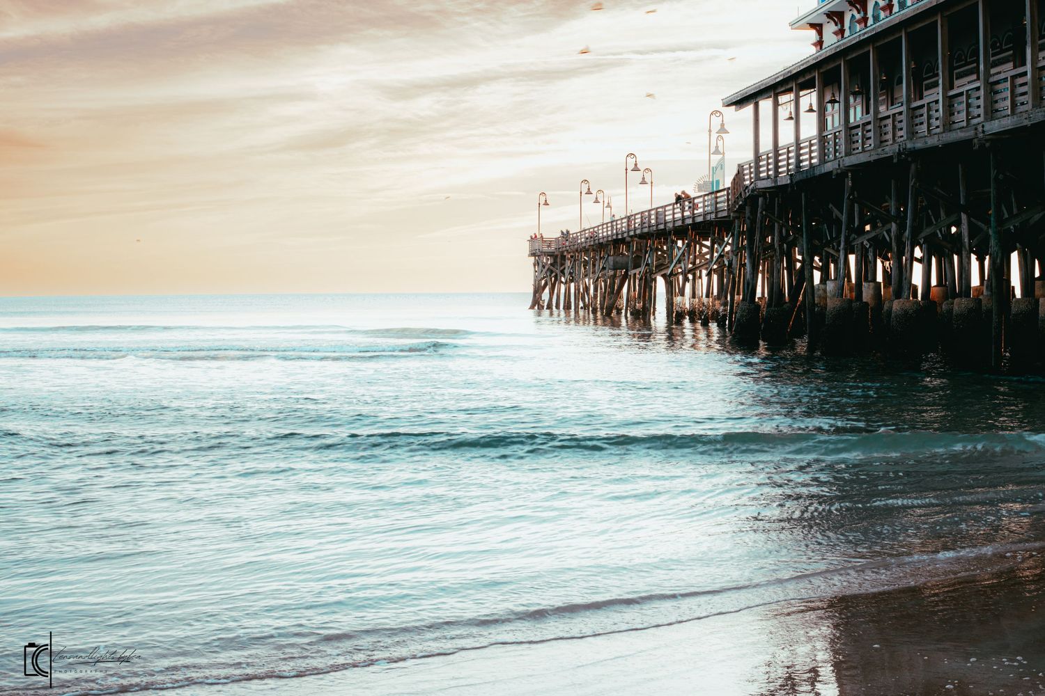 Wooden pier stretches into the ocean at sunset with gentle waves rolling onto the sandy beach.