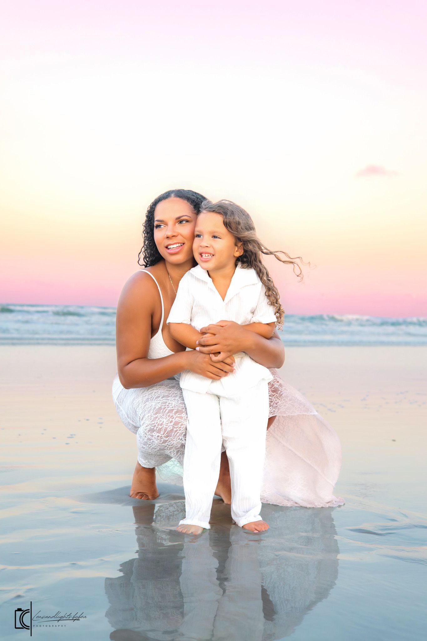A family in white outfits plays and laughs together on a beach at sunset with soft pink sky in the background.