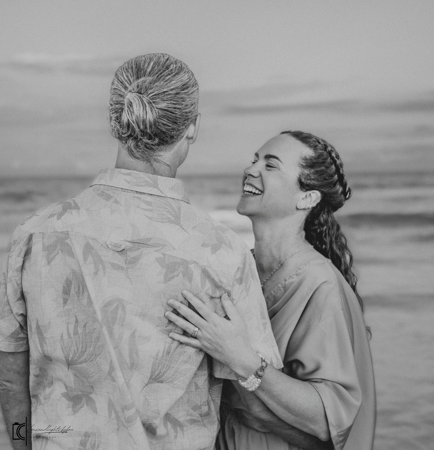 A couple shares a joyful embrace at the beach, laughing together in black and white vintage style photography.