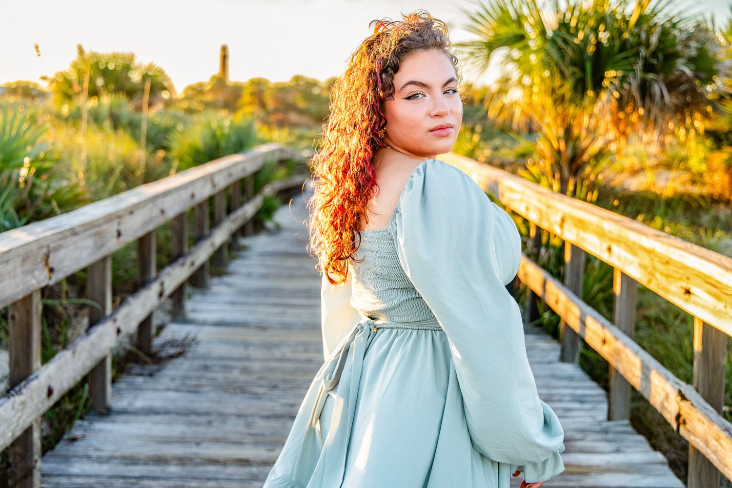 Woman in a sage green dress with curly red hair looks over her shoulder on a wooden beach boardwalk surrounded by palms at golden hour.