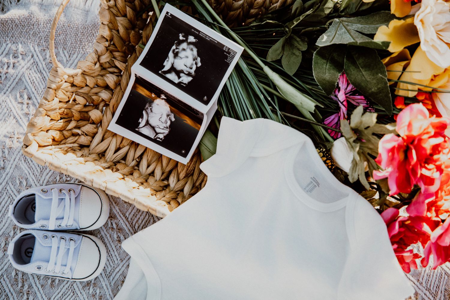 Baby shower flat lay with ultrasound photos, tiny sneakers, white onesie, and colorful flowers in wicker basket.