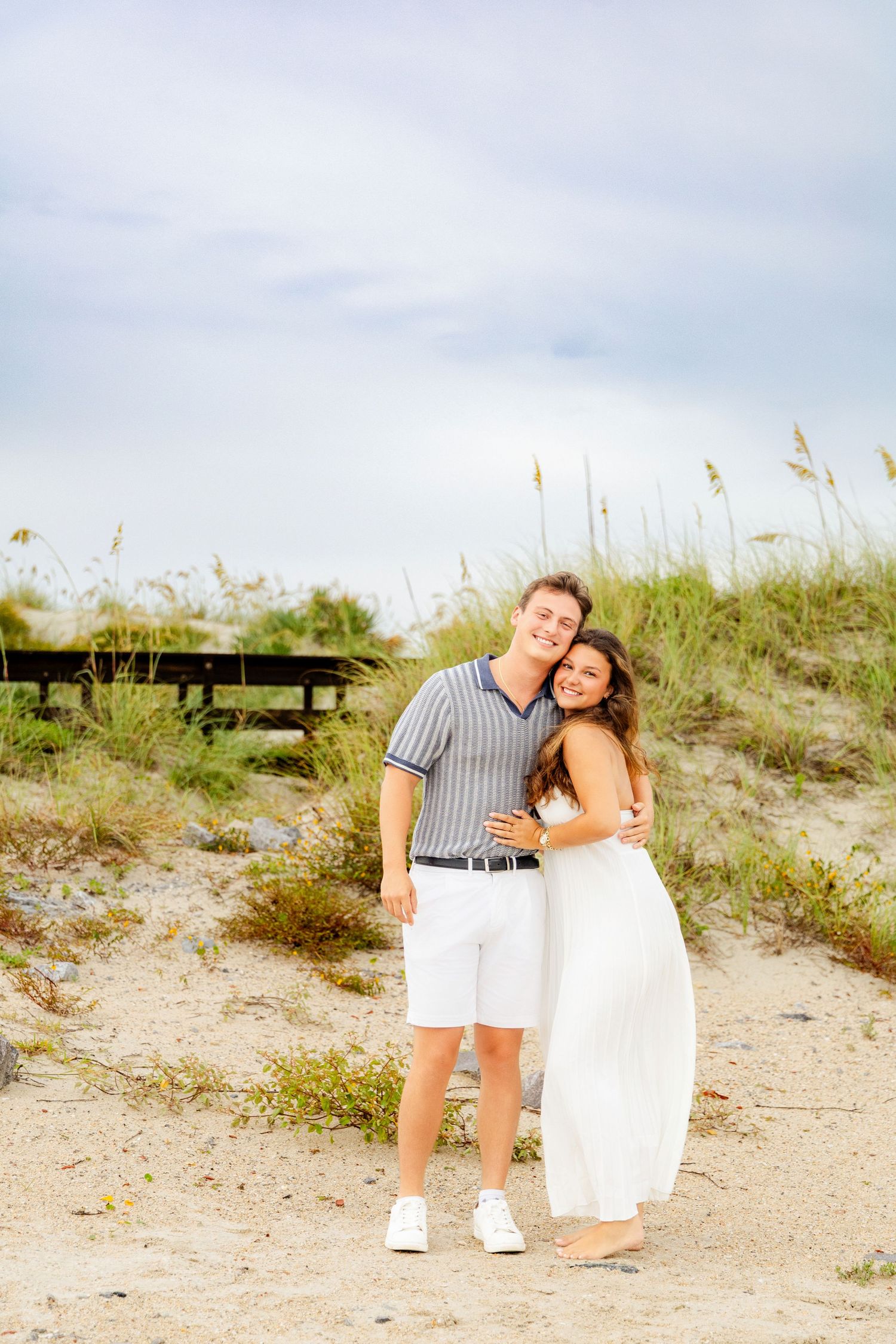 A young couple embracing and smiling on a sandy beach path with sea oats and a cloudy sky behind them.