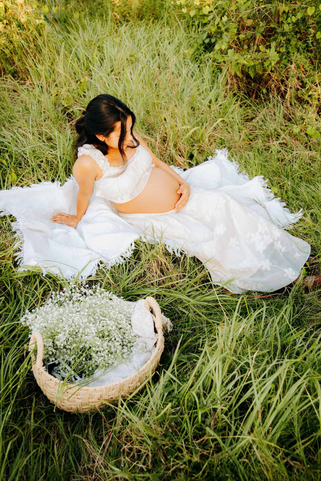 Pregnant woman in white lace dress sitting in green grass with basket of baby's breath flowers.