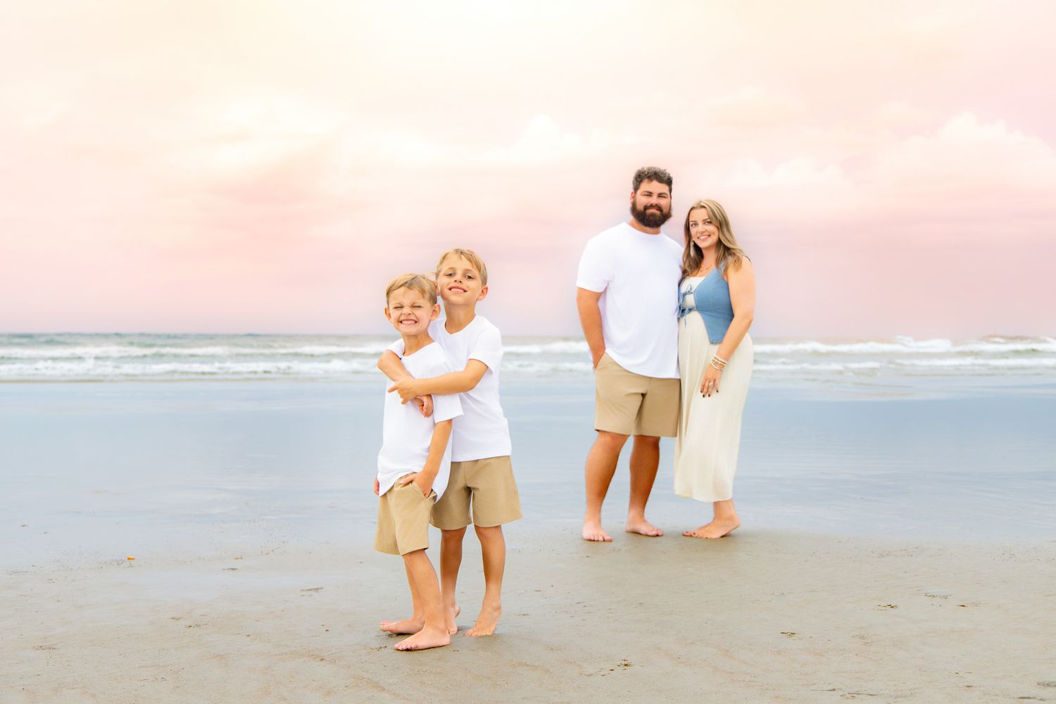 Family of four posing on a beach at sunset, two boys hugging in front while parents stand together behind them.