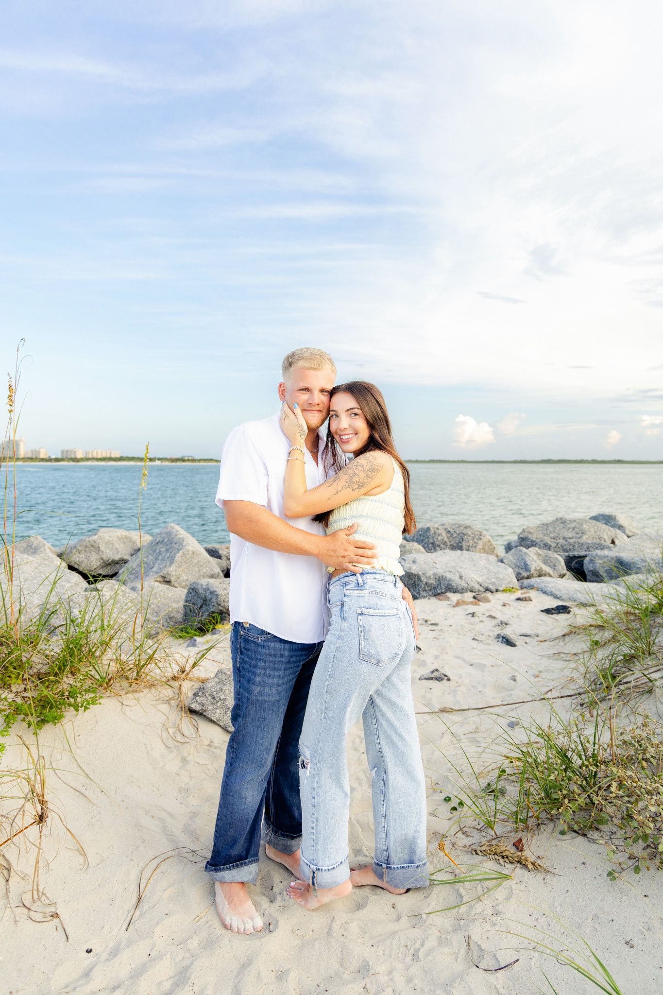 Couple embracing on a sandy beach with rocky shoreline and blue sky in the background.