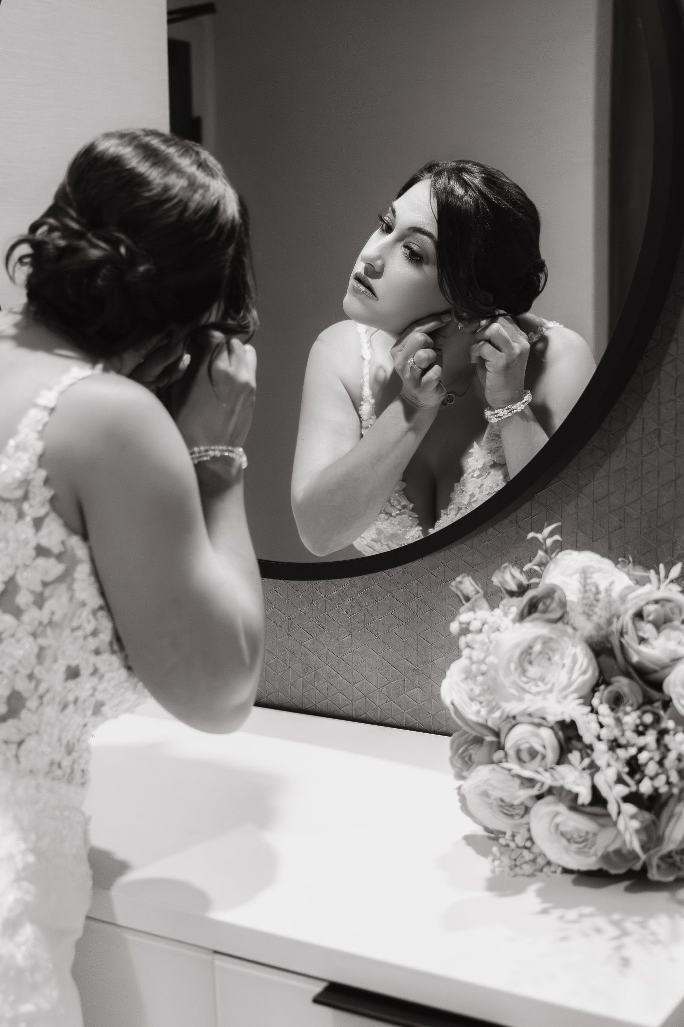 A bride in white adjusts her hair and makeup while looking in a mirror, with white flowers visible in the foreground.