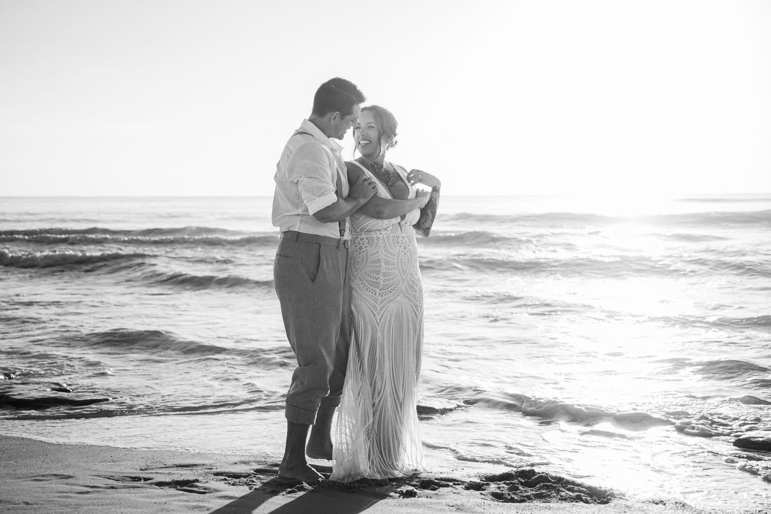 Black and white beach wedding photo of couple embracing at sunset by ocean waves.