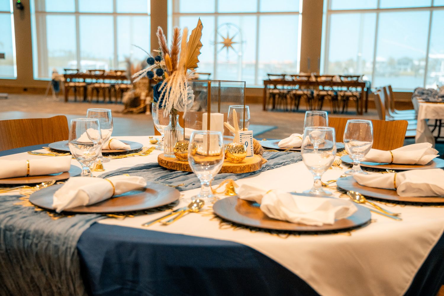Elegant banquet table set with white napkins, gold accents, wine glasses, and floral centerpiece in a bright waterfront venue.