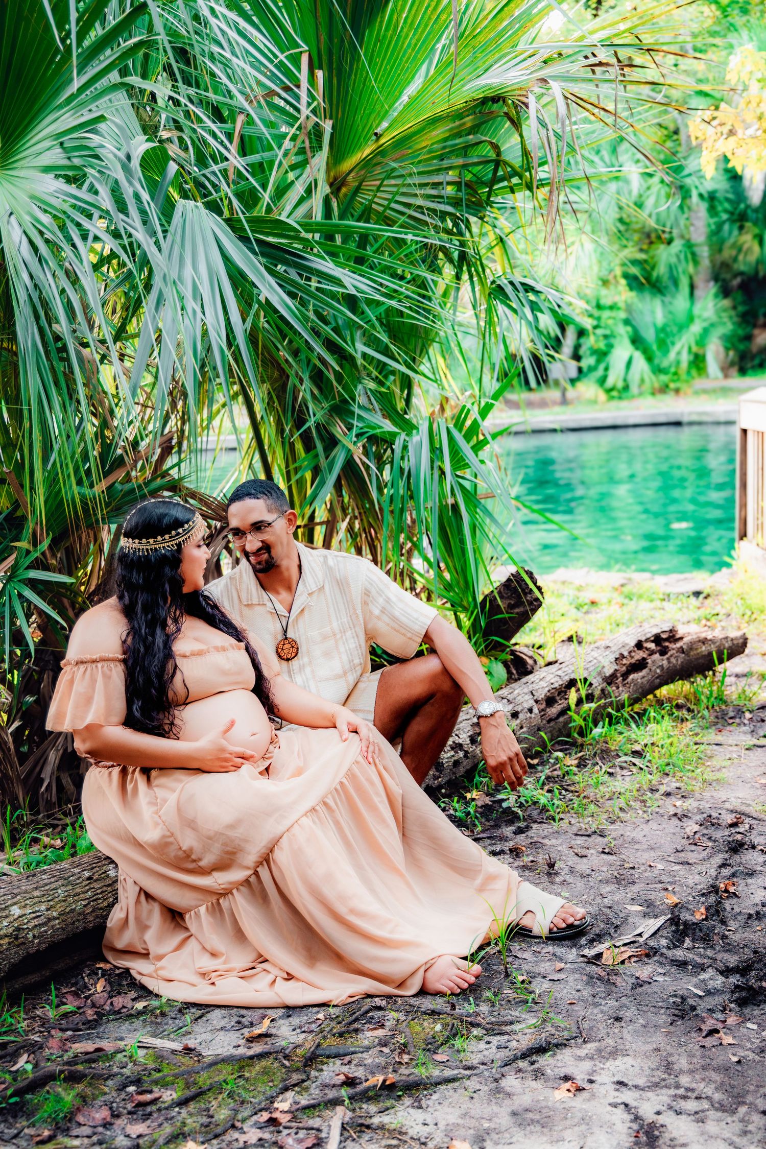 Pregnant couple sitting together under tropical palm trees near turquoise water, woman in peach maternity dress, bohemian style.