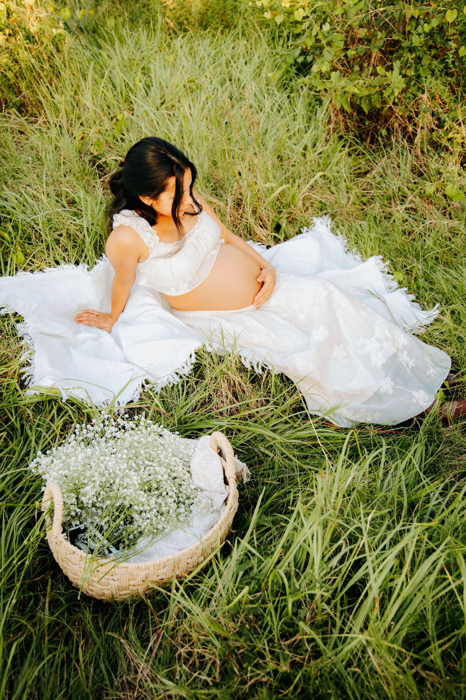 Pregnant woman in white lace dress sitting in green grass with wicker basket of baby's breath flowers.