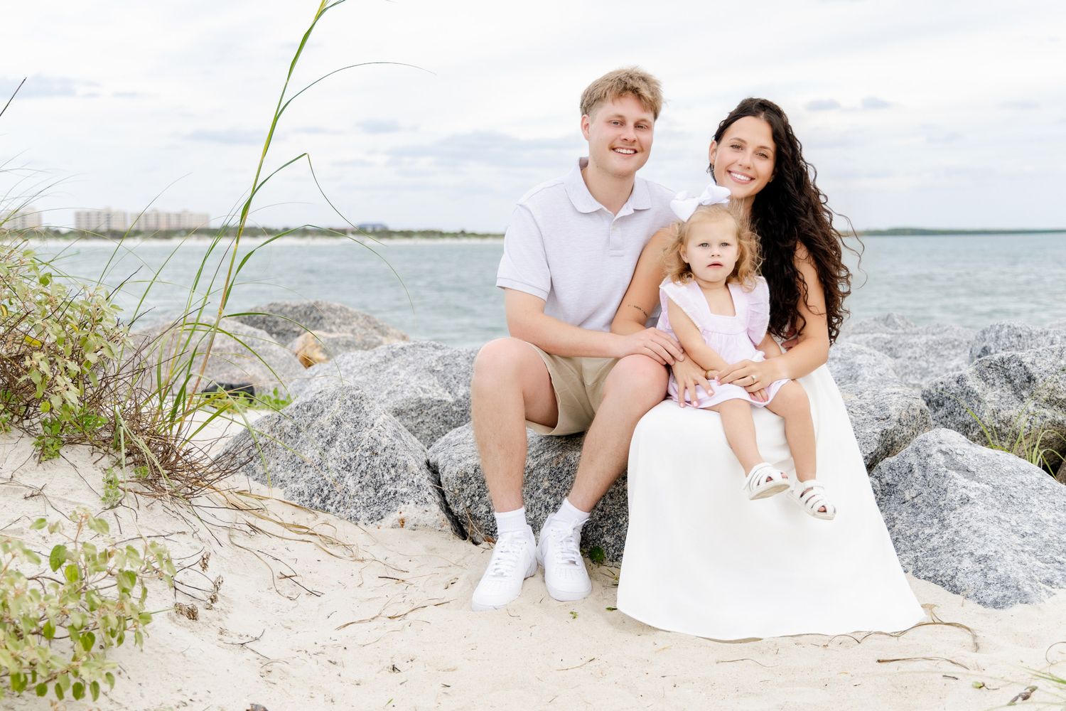 A happy family of three poses on sandy rocks by the ocean, with a young couple and their toddler daughter smiling outdoors.