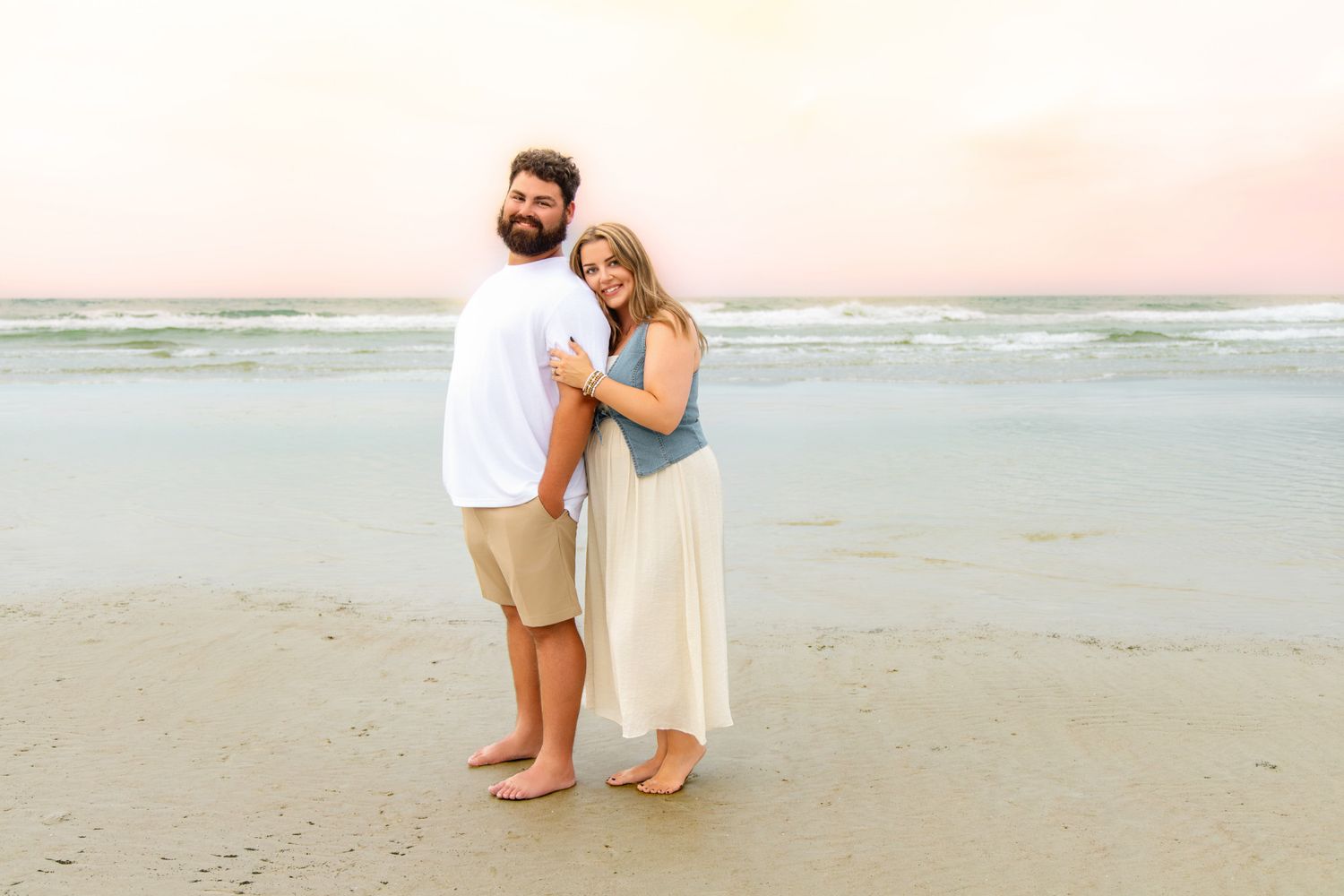 Couple embracing on a sandy beach at sunset, woman in white skirt and denim top, man in white shirt and khaki shorts.