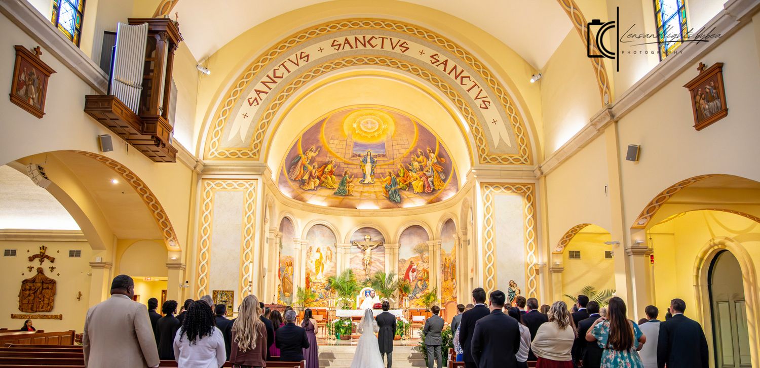 Elegant Catholic church wedding ceremony with ornate golden altar, mosaic artwork, and guests gathered beneath arched ceilings.