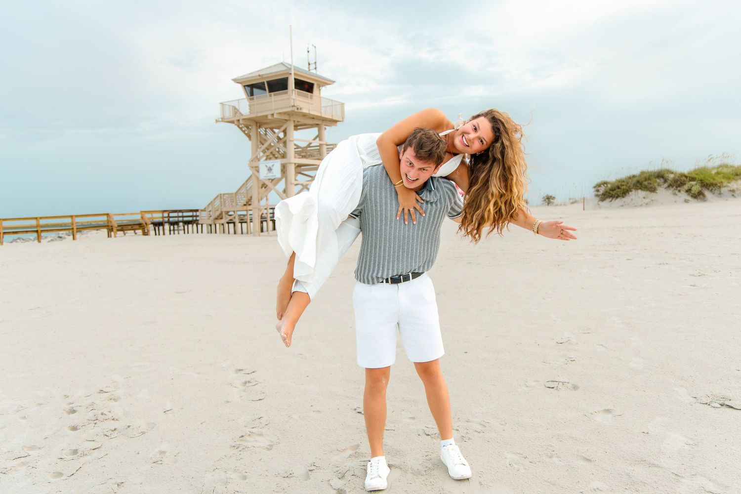 Man giving woman a piggyback ride on a sandy beach with a lifeguard tower in the background.