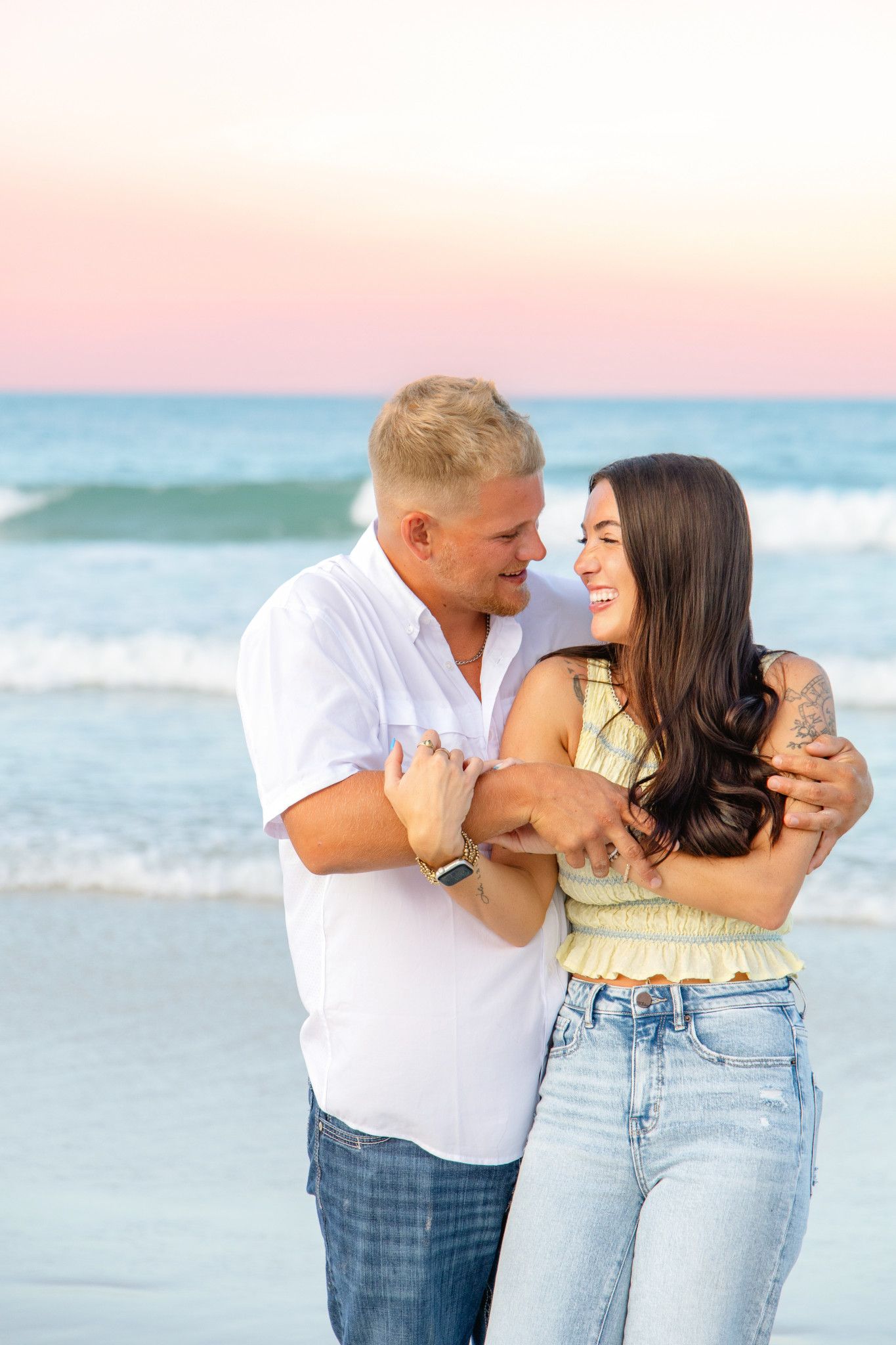 Happy couple embracing and laughing together on a beach at sunset with ocean waves behind them.