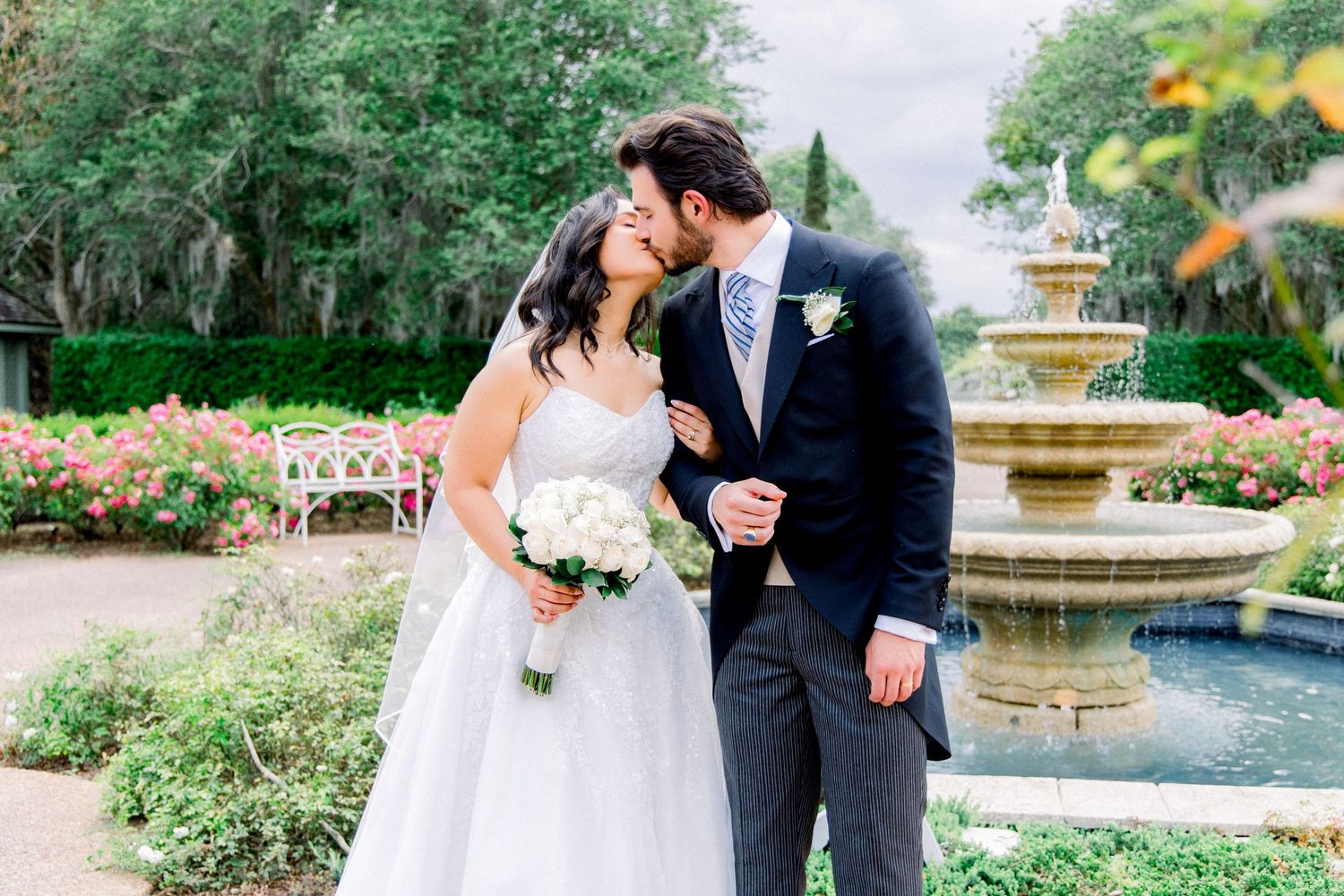 Bride and groom share a kiss near a stone fountain surrounded by lush greenery and blooming roses at an outdoor wedding.
